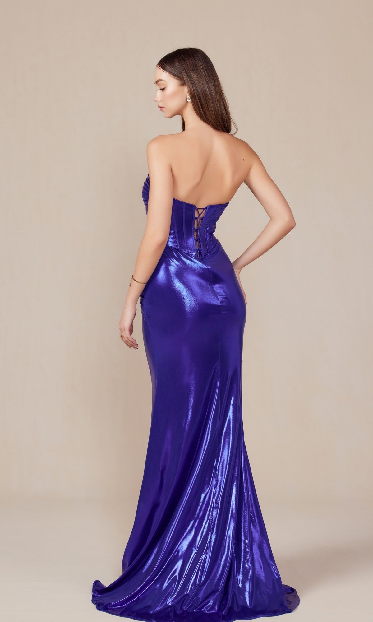 Woman wearing the Pleated Metallic Long Prom Dress (Nox Anabel T1499), featuring a shiny purple finish and lace-up corset back, stands against a plain light background, facing away from the camera.